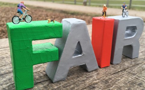 Auf bunten Buchstaben des Wortes Fair stehen Miniaturfiguren